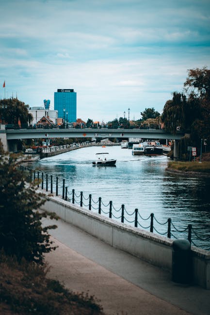 A riverside scene showing a paved walkway with a black chain-link fence along the water's edge, adjacent to a clean concrete surface. The River Thames flows calmly beneath a modern bridge, with a small boat visible on the water. In the background, there are trees, a mix of modern and traditional buildings, and a blue office tower under a cloudy sky. The area appears well-maintained and free of debris, reflecting a tidy and hygienic environment, consistent with professional surface cleaning and maintenance services provided by Carpet Cleaning Putney near Putney Bridge & Riverside.