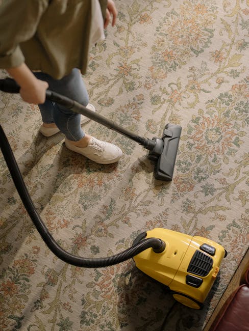 A person operating a yellow vacuum cleaner on a patterned, traditional-style carpet in a room with warm lighting. The individual, wearing light-colored shoes and jeans, is holding the vacuum's wand and is in the process of deep cleaning or surface cleaning the area. The carpet features floral motifs in muted tones, and the room appears tidy and well-maintained. The scene highlights professional domestic cleaning, with Carpet Cleaning Putney providing thorough hygienic maintenance for the space.