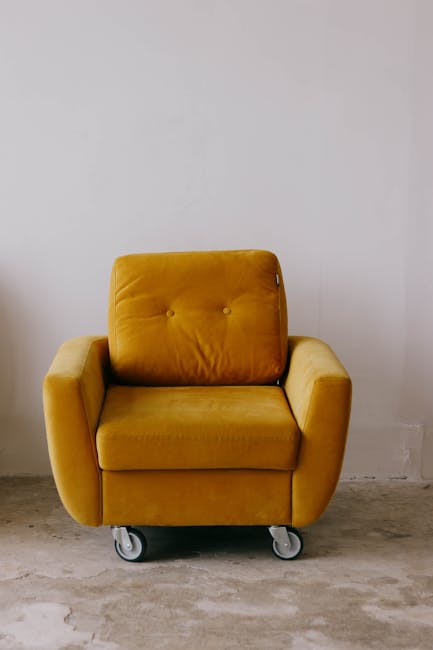 A mustard yellow upholstered armchair with a plush cushion backrest and wide armrests, placed on a concrete floor in front of a plain white wall. The chair has four visible caster wheels underneath, indicating it is mobile and potentially part of a modern, flexible living space or office setting. The surface appears clean and well-maintained, with no visible dust or stains. The lighting is even, suggesting natural or soft artificial light in the room. This image exemplifies domestic or commercial furniture cleaning and maintenance, as recommended by Carpet Cleaning Putney's upholstery cleaning services for landlords to ensure compliance with cleanliness standards.