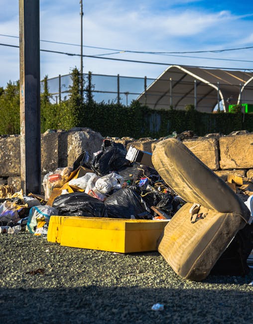 An outdoor scene showing a pile of bulky waste and discarded items, including black trash bags, a yellow plastic container, an old beige car tire, and assorted debris, situated on a gravel surface adjacent to a stone wall. In the background, there is a metal fence, some greenery, and a shed with a curved roof under a partly cloudy sky. The area appears untidy and in need of proper waste disposal and surface cleaning, highlighting the importance of regular hygiene and maintenance. This image demonstrates the type of waste that [COMPANY_NAME], based in Putney, can help remove as part of their domestic cleaning and waste disposal services, ensuring cleanliness and environmental responsibility.