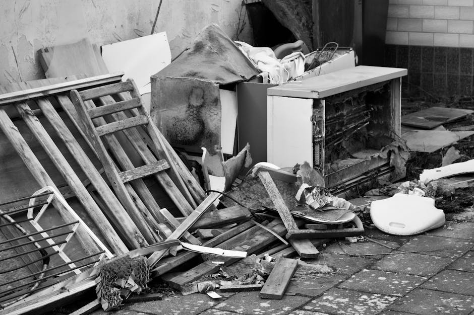 A cluttered outdoor area featuring discarded bulky waste and debris, including wooden pallets leaning against a wall, a small white cabinet with one door open, and various broken pieces of wood and plastic scattered on a paved surface. The scene appears dirty and neglected, with visible grime and dirt on the surfaces. The lighting is natural, highlighting the untidy state of the area, which needs cleaning and proper disposal. The image emphasizes the importance of efficient waste removal and surface cleaning, as promoted by Carpet Cleaning Putney, to maintain a hygienic and tidy environment in residential or commercial spaces.