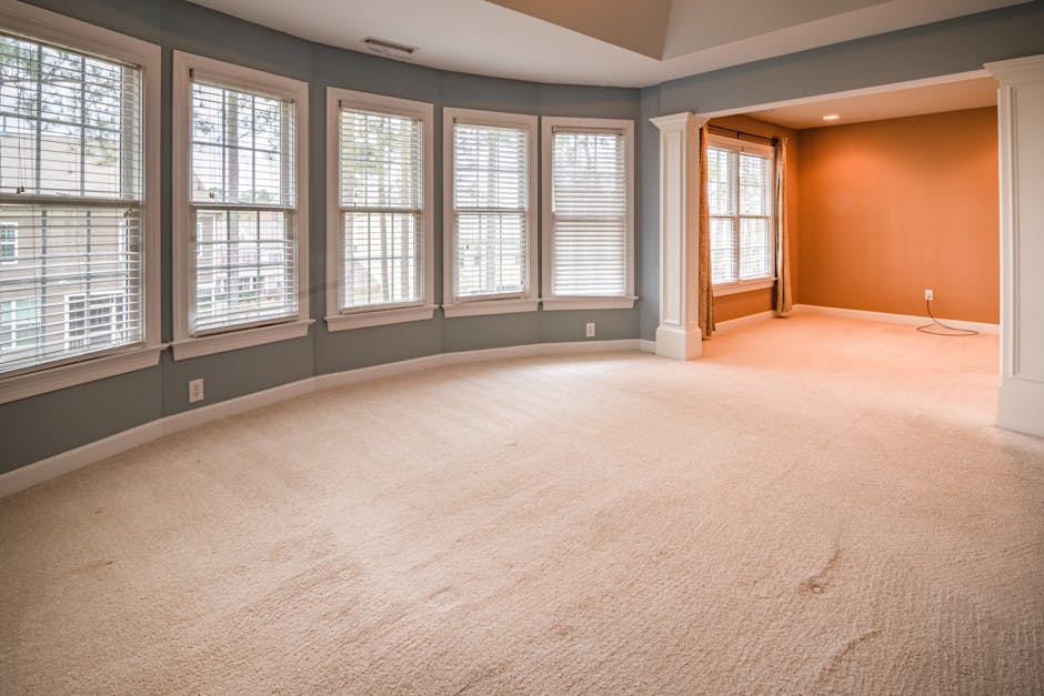 A spacious residential living room with a curved wall of large, white-framed double-hung windows fitted with white blinds, allowing natural light to brighten the beige carpeting and soft gray walls. T