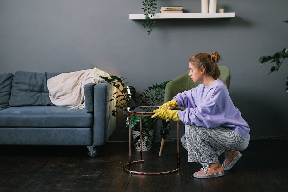 A woman with light brown hair tied in a bun, wearing a lavender sweatshirt, grey and white checkered pants, yellow rubber gloves, and sneakers, is kneeling on a dark wooden floor while cleaning a small round copper-colored side table in a living room. The room features a grey wall with a white shelf holding books and decorative items above, and a grey fabric sofa with a beige blanket draped over one armrest. Next to the woman, there is a potted plant with green leaves placed on the floor. The scene is well-lit, emphasizing the cleanliness and tidy arrangement of the space, highlighting surface cleaning and domestic maintenance activities. Carpet Cleaning Putney is conducted to ensure hygiene and proper maintenance of the home environment, fitting the context of a detailed guide on flats cleaning in SW15 Putney.
