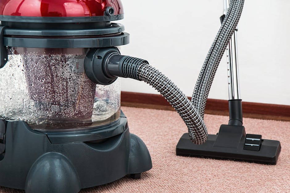 Close-up image of a vacuum cleaner with a transparent dust container filled with debris, connected to a flexible hose and floor tool. The vacuum is placed on a pink carpet in a domestic room with a white wall and wooden skirting board visible in the background. The floor appears clean and well-maintained, indicative of surface cleaning or deep cleaning, emphasizing hygiene and maintenance. The vacuum cleaner is branded with the company's name, Carpet Cleaning Putney, suggesting professional cleaning service.