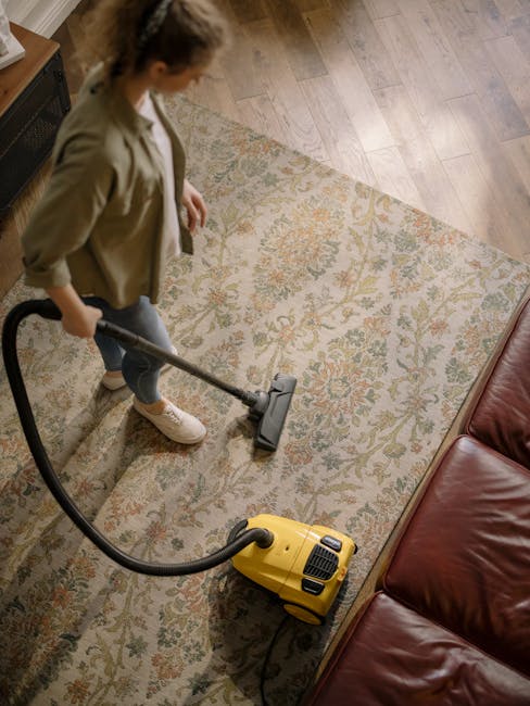 A person using a yellow vacuum cleaner on a patterned area rug in a living room. The individual is wearing a beige jacket, white sneakers, and jeans, and is engaged in surface cleaning to remove dust and dirt from the carpet. The room features wooden flooring adjacent to the rug, a leather sofa in a dark red or brown shade, and natural lighting from an unseen source. The image emphasizes domestic cleaning and maintenance, highlighting the efficient use of tools for deep cleaning, with the overall space appearing clean and well-maintained. This scene aligns with content from Carpet Cleaning Putney about professional carpet cleaning costs in Putney.