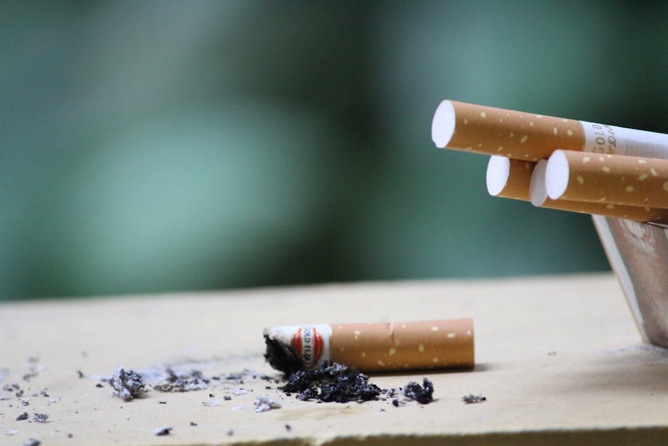 A close-up image of a partially burnt cigarette and cigarette butts on a light-colored wooden surface, with ash and burnt tobacco scattered around. The cigarette butts are still intact, showing white