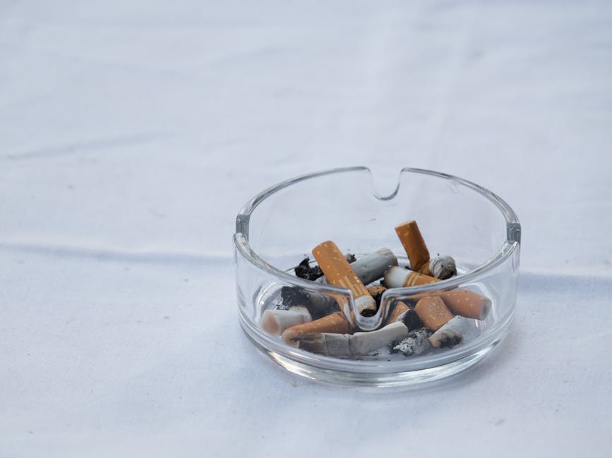 A close-up image of a partially burnt cigarette and cigarette butts on a light-colored wooden surface, with ash and burnt tobacco scattered around. The cigarette butts are still intact, showing white