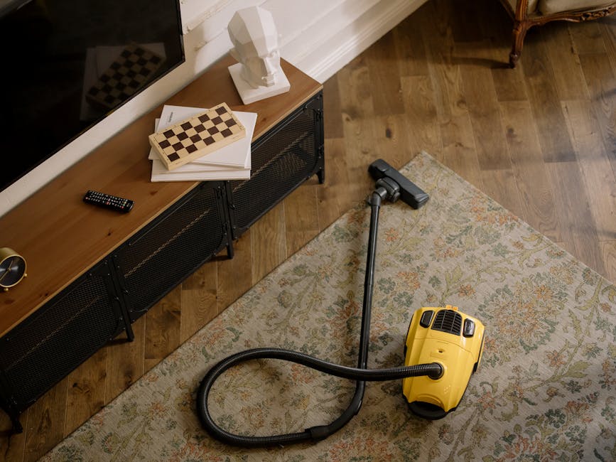 In a living room featuring a hardwood floor, a yellow vacuum cleaner is positioned on a patterned area rug, ready for surface cleaning and deep cleaning. To the left, a modern wooden console table with black metal legs holds a remote control, a chessboard, a white bust sculpture, and a stack of papers. The lighting is natural, illuminating the clean, well-maintained surfaces, with the room exuding a tidy and hygienic atmosphere typical of professional domestic cleaning provided by Carpet Cleaning Putney.