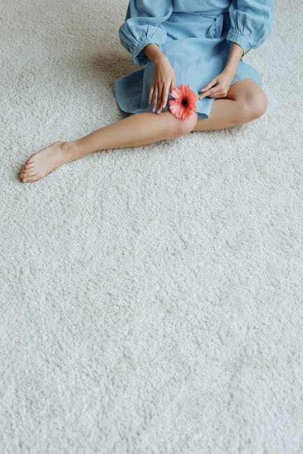 A person dressed in a light blue garment is sitting on a plush, white carpeted floor in a well-lit room. The individual is barefoot, with one leg extended and the other bent, holding a pink flower in one hand while the other hand rests on the carpet. The soft, clean surface of the carpet is free of stains or debris, highlighting a high standard of surface cleaning and hygiene. Visible in the background are no additional objects or furniture, emphasizing the focus on the carpet's cleanliness. The scene illustrates the importance of maintaining spotless carpets as part of domestic cleaning services, such as those offered by Carpet Cleaning Putney, for immediate, professional stain removal and surface care.