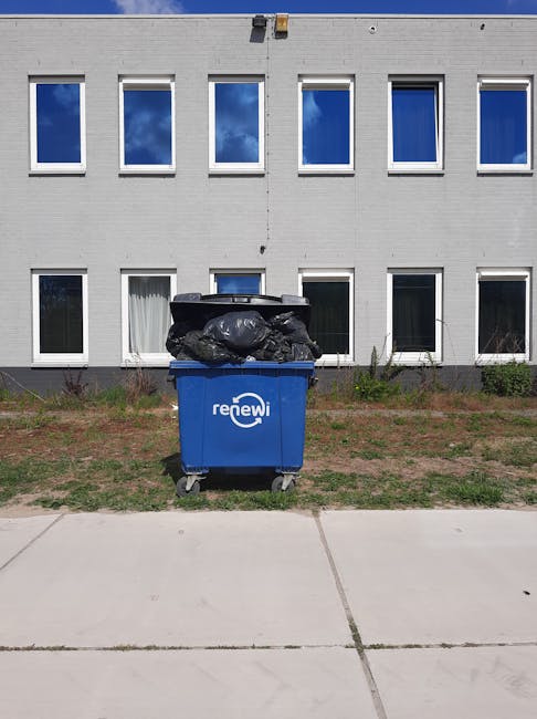 A blue recycling bin labeled 'renewi' positioned on a concrete sidewalk outside a white multi-story building with large rectangular windows. The bin is filled with black garbage bags and some loose waste. The building's exterior is made of white cladding, and the windows have white frames. The scene is illuminated by natural daylight, showcasing a tidy environment with some small plants and grass around the base of the building. This setting illustrates waste disposal practices relevant to Wandsworth Council rules for carpet waste and recycling, as part of domestic surface cleaning and waste management processes discussed by Carpet Cleaning Putney.