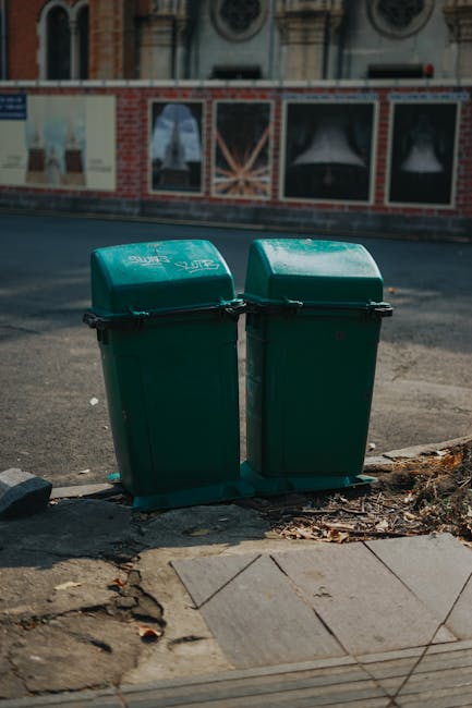 A row of large outdoor waste bins, including black paper recycling bins, white general waste bins, and a large dark container, positioned on a paved surface in front of dense trees and foliage. The bins are securely lidded, with some labels indicating waste types such as 'PAPER' and 'FIBERBOARD ONLY'. The area appears clean and well-organized, with natural lighting highlighting the surfaces. This scene reflects typical waste management and surface cleaning practices associated with domestic cleaning and waste disposal regulations outlined by Wandsworth Council, as covered by Carpet Cleaning Putney in their guidance on carpet waste and recycling rules.